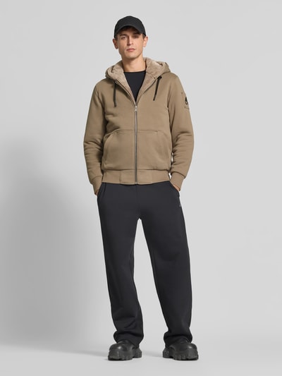 Männermodel trägt Sweatjacke aus Baumwoll-Mix von Moose Knuckles in Beige mit Plüschfutter, Kängurutasche und Reißverschluss, Ganzkörperansicht von vorne.