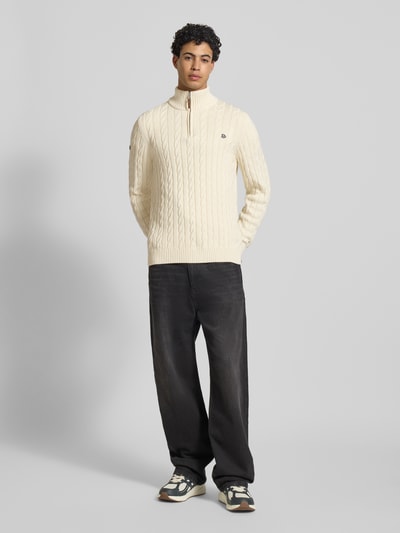 Männermodel trägt Strickpullover aus reiner Baumwolle von Superdry in Creme mit Zopfmuster, Troyer-Kragen, gerippte Abschlüsse, Ganzkörperansicht von vorne.