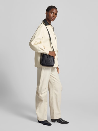 Frauenmodel trägt Crossbody Bag mit Logo-Detail von Tory Burch, schwarzes Strukturmuster, Hauptfach mit Druckknopfverschluss, geteiltes Hauptfach, verstellbarer Schulterriemen, Ganzkörperansicht von vorne.