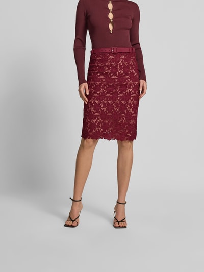 Frauenmodel trägt Slim Fit Rock mit Allover-Spitzenbesatz von Self-Portrait in Weinrot, Knielänge, abnehmbarer Gürtel, blickdichter Unterrock, Ganzkörperansicht von vorne.