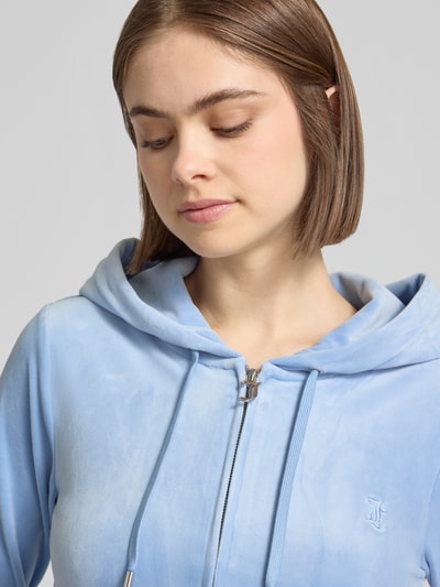 Frauenmodel trägt Slim Fit Sweatjacke aus Samt Modell 'Robertson' von Juicy Couture in Hellblau mit Kapuze, Tunnelzug, Reißverschluss, geteilte Känguru-Tasche, Elastischer-Material-Mix, elastische Abschlüsse; Nahaufnahme Oberkörper.