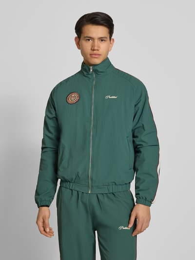 Männermodel trägt Trackjacket mit Label-Stitching Modell 'Slipstream' von PROHIBITED in Dunkelgrün, Reißverschluss, Stehkragen, Ganzkörperansicht von vorne, Relaxed Fit, schlichte glatte Textur.