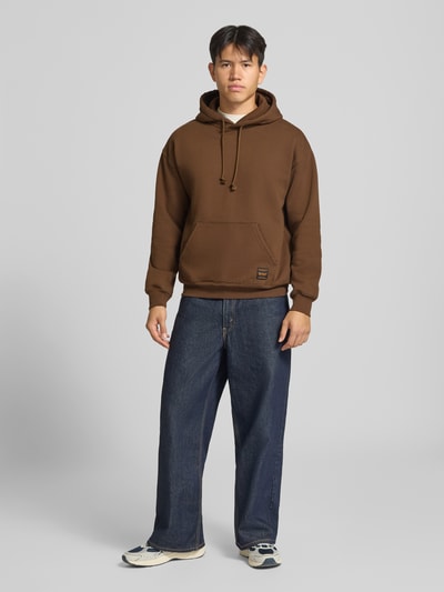 Männermodel trägt Regular Fit Hoodie aus Baumwoll-Mix von Levi's® in Braun mit Kängurutasche, kombiniert mit Jeans, Ganzkörperansicht von vorne.