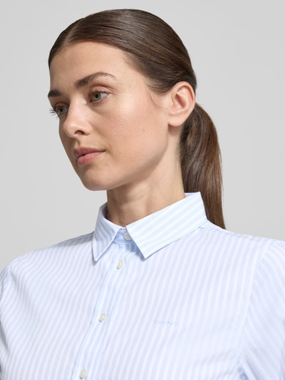 Kobieta modelka nosi Regular Fit Bluse mit Streifenmuster und Label-Stitching od Gant, czysta bawełna, krój regular fit, niebiesko-białe paski, kołnierzyk koszulowy, detal z logo, zbliżenie na górną część ciała.