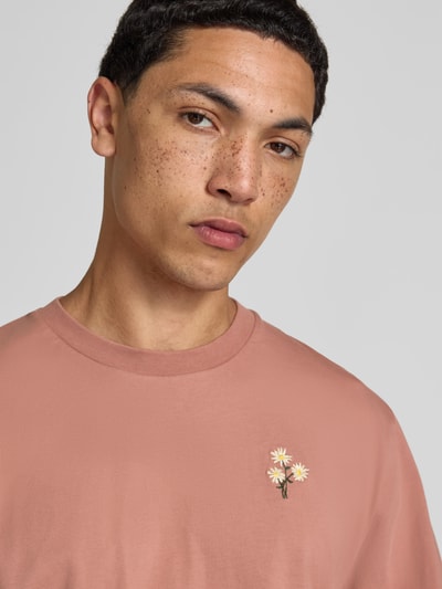Männermodel trägt T-Shirt mit Rundhalsausschnitt von Forét in Rosa, Reine Baumwolle, Regular-Fit, florales Stitching, Nahaufnahme Oberkörper.