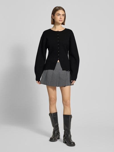 Frauenmodel trägt Regular Fit Strickjacke aus Viskose-Mix in Strick-Optik von Gina Tricot, schwarz, Rundhalsausschnitt, durchgehende Knopfleiste, elastische Ärmelabschlüsse, kombiniert mit kurzem grauem Rock, Ganzkörperansicht von vorne.