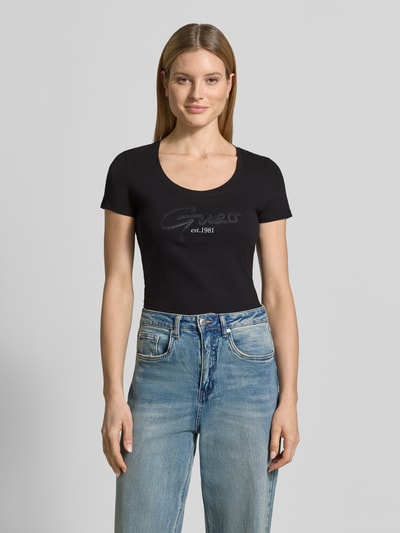 Frauenmodel trägt Damen T-Shirt mit U-Ausschnitt Modell 'SCRIPT' von Guess aus Baumwoll-Elasthan-Mix, körpernaher Schnitt, Label-Stitching, schwarze Farbe, Rückenlänge bei Größe S: 61 cm, Ganzkörperansicht von vorne.