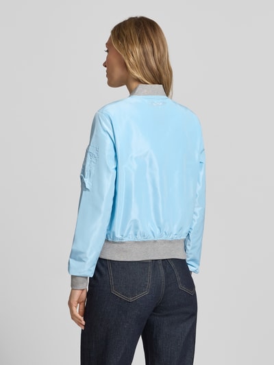 Frauenmodel trägt hellblaue Bomberjacke mit Label-Patch und Reißverschlusstasche am Ärmel von Original Bombers, Rückenansicht, glatte Textur, lockerer Schnitt, Stehkragen, Logo-Stitching, Druckknopfverschluss an seitlichen Eingrifftaschen, elastische Abschlüsse, Rückenlänge bei Größe S 53 cm, Schulterbreite 37 cm, Brustweite 48 cm, Ärmellänge 63 cm. Ganzkörperansicht Rückansicht.