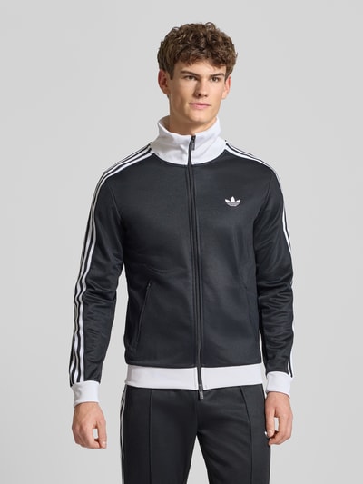 Männermodel trägt Regular Fit Trainingsjacke mit Stehkragen Modell 'Classic' von adidas Originals im Baumwoll-Mix, Schwarz mit weißen Details, Zweiwege-Reißverschluss, Logo-Stitching, elastische Abschlüsse, Rückenlänge 68 cm, Ärmellänge 65 cm, Ganzkörperansicht von vorne.