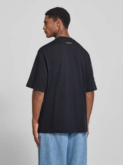 Männermodel trägt Comfort Fit T-Shirt mit Motiv-Print von Armani Exchange in Schwarz, Allover-Look, Rundhalsausschnitt, Rückenlänge 76 cm, Ganzkörperansicht Rückansicht.