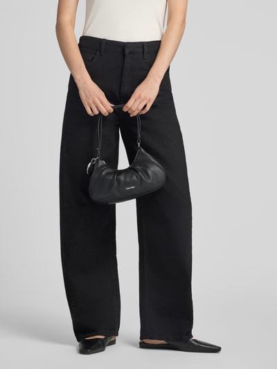 Frauenmodel trägt schwarze Henkeltasche mit Label-Prägung Modell 'ENGRAVED HW SMALL BAG' von CK Calvin Klein, Damen Henkeltasche, Ganzkörperansicht von vorne.