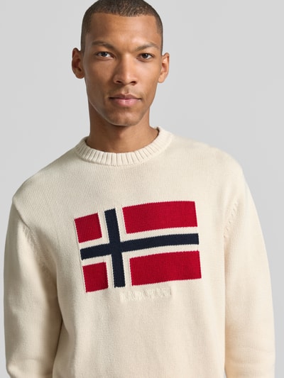 Männermodel trägt Strickpullover aus reiner Baumwolle Modell 'MURA' von Napapijri in Beige mit Norwegenflagge, Nahaufnahme Oberkörper.