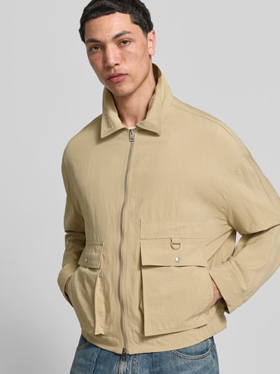 Männermodel trägt Regular Fit Jacke mit Umlegekragen von Maison Kitsuné in Beige, aufgesetzte Taschen, Zweiwege-Reißverschluss, Sportmanschetten, Ganzkörperansicht von vorne.