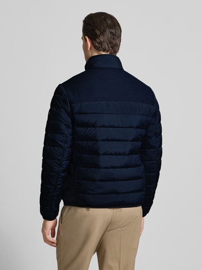 Męski model nosi Regular Fit Steppjacke mit seitlichen Stretcheinsätzen od Tom Tailor, kolor granatowy, krój regular fit, elastyczne wstawki boczne, długie rękawy, stójka, tylny karczek, widoczna naszywka z logo, widok całej sylwetki z tyłu.