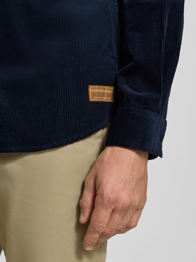 Mężczyznamodel nosi Regular Fit Cordhemd mit Brusttasche od Scotch & Soda z bawełny, naszywka z logo, krój regular fit, brustasza, Kołnierzyk typu kent, długość tyłu w rozmiarze M: 78 cm, szerokość ramion w rozmiarze M: 46 cm, długość rękawów w rozmiarze M: 66 cm, kolor granatowy, beżowe spodnie, detal z bliska na dolną część koszuli i rękaw, z boku.