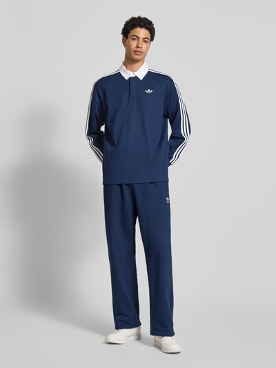 Männermodel trägt Poloshirt mit Label-Stitching von adidas Originals in Dunkelblau mit weißen Details, reine Baumwolle, Ganzkörperansicht von vorne.