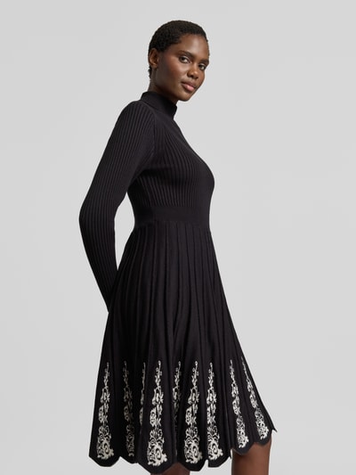 Modelka nosi knielanges Strickkleid aus Viskose-Mix von Apricot, czarny kolor, hafty kwiatowe, Efekt prążkowania, taliowany krój, stójka, długi rękaw, spódnica linia a z plisowaniem, długość do kolan, długość tyłu w rozmiarze S: 99 cm, szerokość w rozmiarze S: 34 cm, długość rękawów w rozmiarze S: 60 cm, zdjęcie całej sylwetki z przodu.