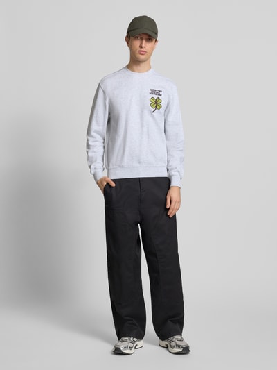 Mężczyzna nosi Sweatshirt mit Label-Detail od Carhartt Work In Progress, szary, czysta bawełna, krój regular fit, ściągacze, dekolt okrągły, długość tyłu w rozmiarze M: 67 cm, długość rękawa: 66 cm, widok całej sylwetki z przodu.