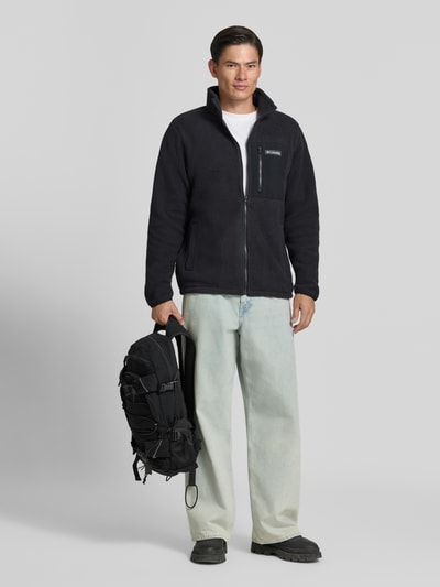 Männermodel trägt schwarze Fleecejacke mit Stehkragen Modell 'Jasper' von Columbia, Struktur weich, Ganzkörperansicht von vorne. Herren Fleecejacke, gerade Passform, durchgehender Reißverschluss, Brusttasche mit Reißverschluss, Label-Patch, seitliche Eingrifftaschen, elastische Ärmelabschlüsse, Rückenlänge 72 cm, Ärmellänge 71 cm, Schulterbreite 47 cm, Brustweite 46 cm.