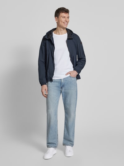 Męski model nosi kurtkę z kapturem model 'NIGLE' od Peuterey, krój regular fit, zamek błyskawiczny dwustronny, wpuszczane kieszenie, długość tyłu w rozmiarze M 69 cm, szerokość ramion w rozmiarze M 47 cm, szerokość klatki piersiowej w rozmiarze M 56 cm, długość rękawów w rozmiarze M 68 cm, kolor granatowy, proste formy, gładka tekstura, całe ciało widoczne, widok z przodu.
