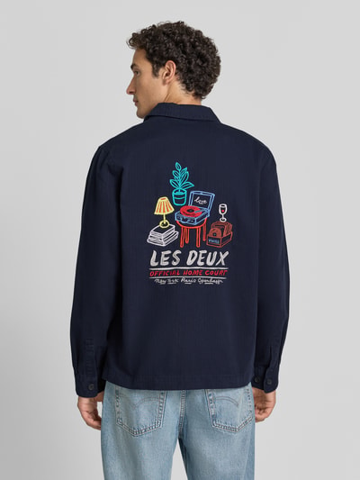 Männermodel trägt Regular Fit Hemdjacke aus reiner Baumwolle von Les Deux in Dunkelblau mit buntem Rückenprint, Ganzkörperansicht Rückansicht. Kontext: Herren Hemdjacke von Les Deux, reine Baumwolle, Denim, Kentkragen, langärmelig, aufgesetzte Brust- und Eingrifftaschen, Label-Stitching, durchgehende Knopfleiste, Rückenlänge bei Größe M 74 cm.