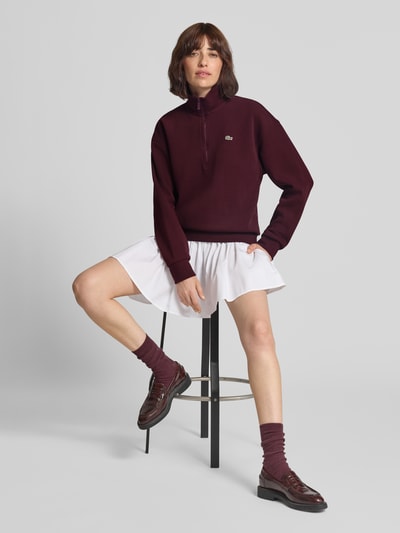 Frauenmodel trägt Relaxed Fit Sweatshirt aus Baumwoll-Mix von Lacoste in Dunkelrot, kombiniert mit weißem Rock, glatter Stoff, Ganzkörperansicht von vorne. Damen Sweatshirt von Lacoste.