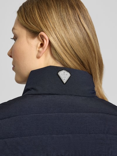 Kobieta nosi kurtkę pikowaną z zamkiem błyskawicznym i kieszeniami zapinanymi na zamek błyskawiczny Steppjacke mit Reißverschlusstaschen Modell 'GS 111' od G.I.G.A. DX, krój slim fit, stójka, długość tyłu w rozmiarze 36: 67 cm, szerokość ramion w rozmiarze 36: 38 cm, szerokość klatki piersiowej w rozmiarze 36: 49 cm, długość rękawów w rozmiarze 36: 65 cm, detaliczny widok tyłu.