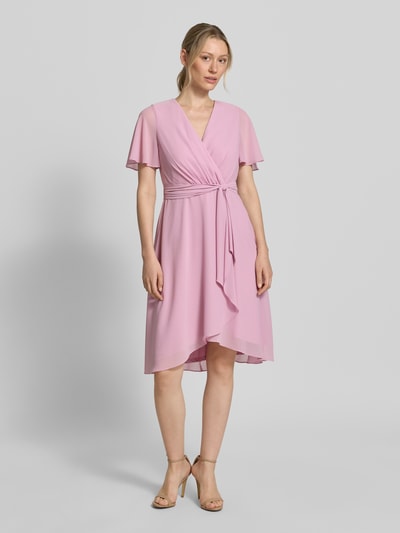 Vrouwenmodel draagt knielanges Cocktailkleid in Wickel-Optik van Christian Berg Cocktail in lichtroze chiffon, A-lijn, V-hals, met bindriem, ruglengte 100 cm, borstwijdte 33 cm, mouwlengte 22 cm, geheel van voren.