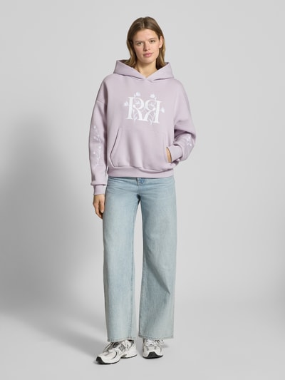 Frauenmodel trägt Relaxed Fit Hoodie mit Logo-Print von Review in Flieder, Känguru-Tasche, florales Stitching, elastische Abschlüsse, Ganzkörperansicht von vorne.
