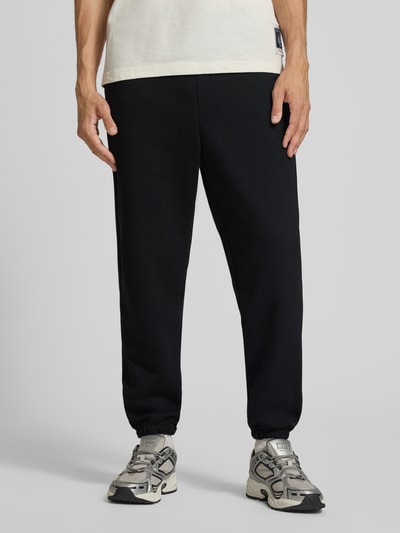 Männermodel trägt Sweatpants mit Label-Print von Alpha Industries in Schwarz, lockere Passform, Baumwoll-Mix, Ganzkörperansicht von vorne.
