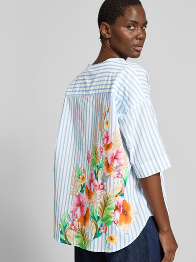 Männermodel trägt Relaxed Fit Bluse mit Motiv-Print von Emily Van den Bergh aus bawełna, dekolt w serek, rękaw o dł. 1/2, długość tyłu 48 cm, długość rękawów 24 cm, kolor biało-niebieski z kwiatowym nadrukiem z motywem na plecach, Ganzkörperansicht Rückansicht.