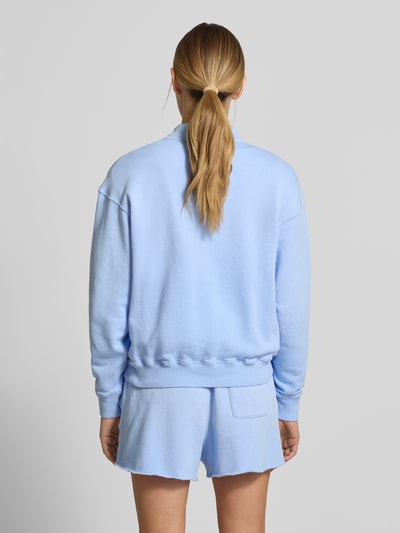 Modelka kobieca nosi Regular Fit Sweatshirt aus Baumwoll-Mix od Polo Ralph Lauren, jasnoniebieski, bawełniany, gładka tekstura, krój regular fit, logo, polokragni z dekoltem V, zapięcie na dwa guziki, długie rękawy, ściągacze na rękawach i dole, widok całej sylwetki z tyłu.