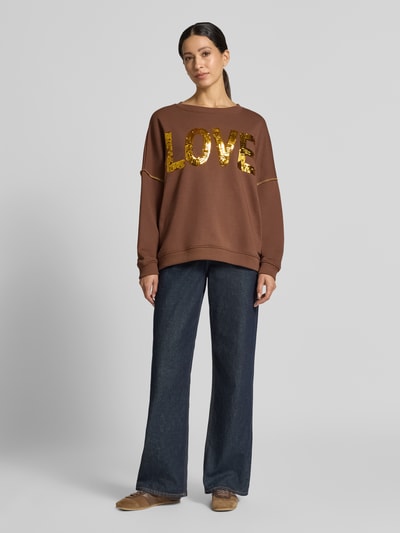 Frauenmodel trägt Oversized Sweatshirt aus reiner Baumwolle Modell 'LOVE' von miss goodlife in Braun mit goldfarbenem Paillettenbesatz, Statement-Print und Rippbündchen, Ganzkörperansicht von vorne.
