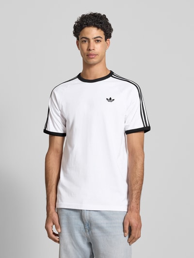 Männermodel trägt T-Shirt mit Label-Stitching von adidas Originals, weiß mit schwarzen Details, Baumwoll-Mix, Regular Fit, Ganzkörperansicht von vorne.