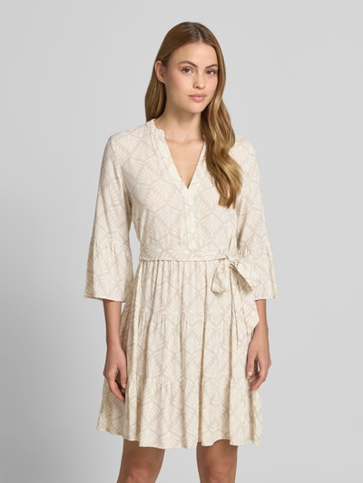 Frauenmodel trägt Tailliertes Tunikakleid aus reiner Viskose Modell 'MELBA' von Vero Moda, Optik:Allover-Muster, Mini-Länge, V-Ausschnitt, Bindegürtel, Ärmelform:Trompetenärmel, Volant-Saum, Material: reine Viskose, Creme mit dezentem Muster, glatte Textur, Ganzkörperansicht von vorne.