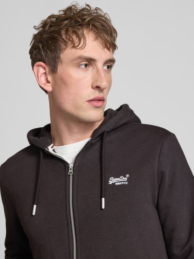 Männermodel trägt Sweatjacke mit Kapuze und Logo-Stitching von Superdry in Dunkelgrau, Baumwoll-Mix, Nahaufnahme Oberkörper, Regular-Fit, Reißverschluss, Kordelzug, Label-Detail, seitliche Eingrifftaschen, gerippte Abschlüsse, Rückenlänge 66 cm, Ärmellänge 62 cm.