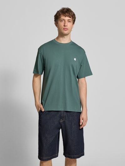 Męski model nosi t-shirt z wyhaftowanym logo, model 'Madison' od Carhartt Work In Progress, bawełna, krój regular fit, okrągły dekolt, długość tyłu w rozmiarze M: 75 cm, widok całej sylwetki z przodu.