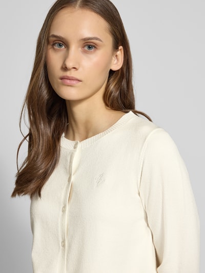 Modelka kobieca ma na sobie Strick-Cardigan aus Viskose-Mix mit langen Ärmeln od Review, w kolorze kremowym, krój regular fit, guziki, haftowane logo, ściągacze na rękawach i u dołu, długość pleców 53 cm, długość rękawa 63 cm, zdjęcie: zbliżenie górnej części ciała.