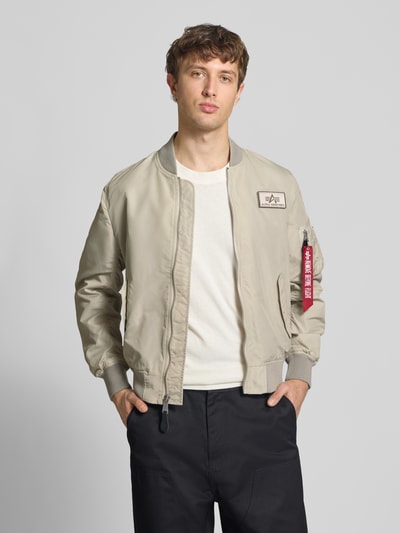 Męskimodel nosi Regular Fit Bomberjacke mit Logo-Patch od Alpha Industries w kolorze beżowym, krój regular fit, stójka, wpuszczane kieszenie, zamek błyskawiczny, kieszeń na rękawie z zamkiem błyskawicznym, długość tyłu w rozmiarze M: 65 cm, szerokość ramion w rozmiarze M: 45 cm, szerokość klatki piersiowej w rozmiarze M: 54 cm, długość rękawów w rozmiarze M: 69 cm; Ganzkörperansicht von vorne.