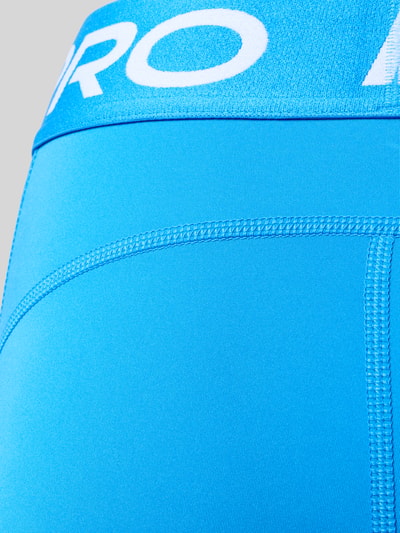 Frauenmodel trägt Leggings mit elastischem Bund und Logo von NIKE TRAINING in leuchtendem Blau, glatte Struktur, Nahaufnahme Hüftbereich.