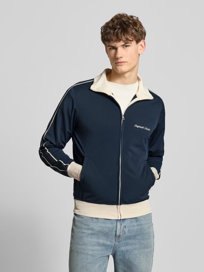Mannenmodel draagt Slim Fit Sweatjacke met logo-stitching model 'Niagara' van Jack & Jones, staande kraag, steekzakken, ritssluiting, elastische boorden, kleur donkerblauw met beige, context: heren sweatjacke, ruglengte maat M 67 cm, mouwlengte maat M 65 cm, voorkant totaalshot