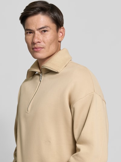 Męski model nosi Regular Fit Sweatshirt aus Baumwoll-Mix mit Troyer-Kragen od DRYKORN, beżowy, wyhaftowane logo, długość tyłu 64 cm, długość rękawów 57 cm, mieszanka bawełny, kołnierz zapinany, krój regular fit, Ganzkörperansicht z przodu.