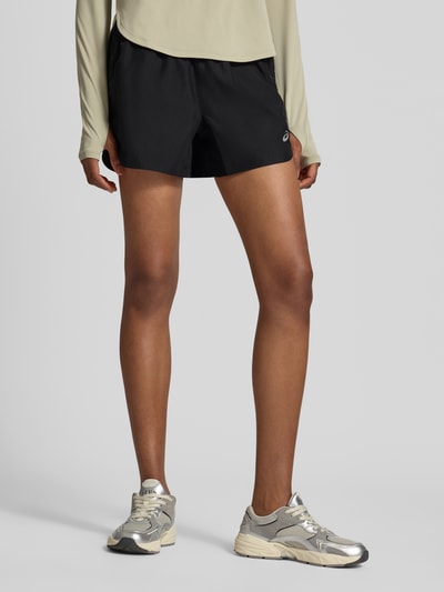 Frauenmodel trägt schwarze Shorts mit elastischem Bund Modell 'NAGINO' von ASICS TIGER, reflektierende Details, seitliche Taschen, Ganzkörperansicht von vorne.