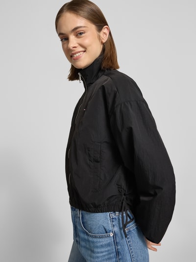 Modelka kobieca nosi czarny bluzon 'Relaxed Fit Blouson mit Taillenbändern' od Tommy Jeans, efekt: jednolity kolor, krój relaxed fit, stójka, zamek błyskawiczny, wyhaftowane logo, tasiemka w talii, długi rękaw, elastyczne zakończenie rękawów, boczne wpuszczane kieszenie na zatrzask; widok całej sylwetki z przodu
