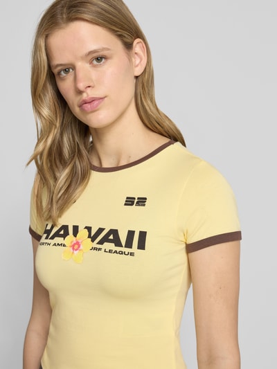 Frauenmodel trägt Slim Fit T-Shirt mit Statement-Print Modell 'Molly' von Noisy May, gelb mit braunen Details, elastischer Baumwoll-Mix, Nahaufnahme Oberkörper.
