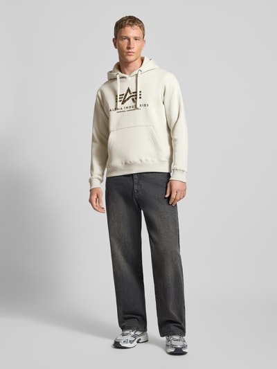 Männermodel trägt Hoodie mit Logo und Känguru-Tasche von Alpha Industries in Beige, Baumwoll-Mix, Regular-Fit, Ganzkörperansicht von vorne.
