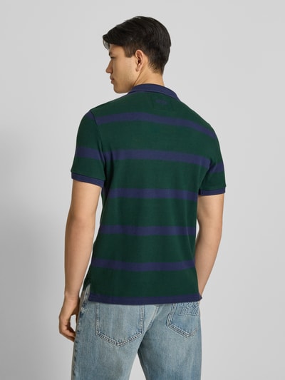 Männermodel trägt Slim Fit Poloshirt mit Label-Stitching und Knopfleiste von Superdry, grün-blau gestreift, Reine Baumwolle, Rückenlänge bei Größe M 70 cm, Ganzkörperansicht Rückansicht.