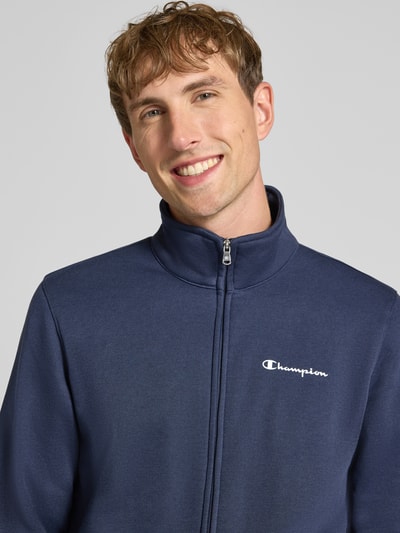 Męskimodel nosi 'Sweatanzug mit Label-Details' od Champion, kolor granatowy, mieszanka bawełny, stójka, detal z logo, zamek błyskawiczny, prosty krój, wpuszczane kieszenie, elastyczny pas, Nahaufnahme górnej części ciała.