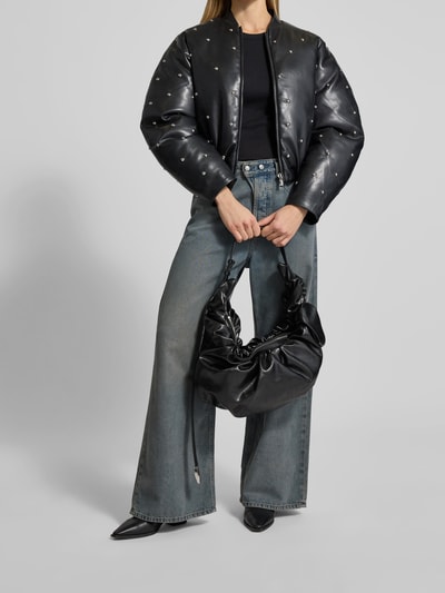Frauenmodel trägt Shoulder Bag mit Raffungen von Diesel in schwarzer Leder-Optik mit verstellbarem Schulterriemen, Hauptfach mit Reißverschluss, Reißverschlussfach innen, 50 cm breit, 29 cm lang, 18 cm tief, Ganzkörperansicht von vorne.