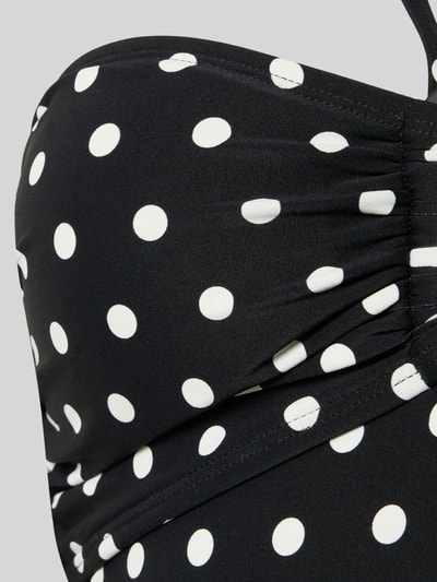 Frauenmodel trägt Bandeau-Badeanzug im Allover-Look Modell 'CLASSIC DOT BANDEAU' von Michael Kors, schwarzer Material-Mix mit Stretch-Anteil und weißen Punkten, körpernahe Passform, Nahaufnahme Oberkörper.
